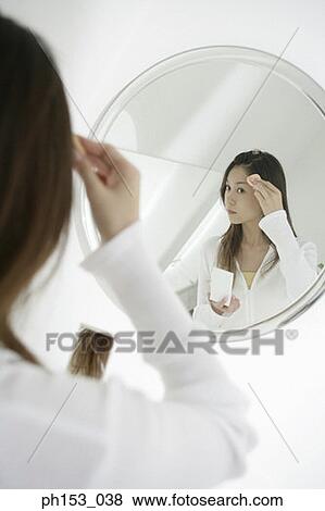 Stock Photo - Reflection of a young woman applying face powder on her forehead in the mirror . Fotosearch