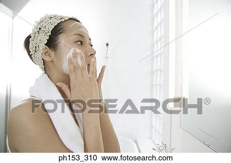 Stock Image - Side profile of a young woman applying soap on her face. Fotosearch