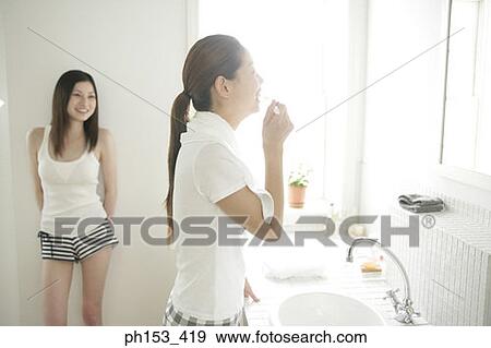 Side profile of a young woman brushing her teeth with another young woman looking at her View Large Photo Image Stock Photo - Side profile of a young woman brushing her teeth with another young woman looking at her. Fotosearch