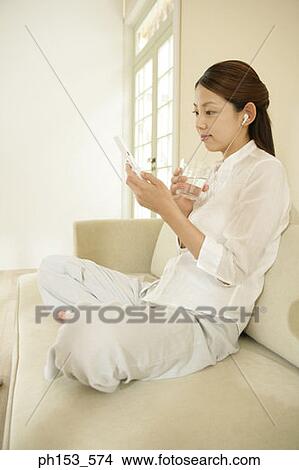 Side profile of a young woman listening to music on a couch and using a mobile phone View Large Photo Image Picture - Side profile of a young woman listening to music on a couch and using a mobile phone. Fotosearch