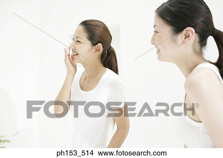 Side profile of a young woman looking at another young woman wiping her face with a cotton pad View Large Photo Image Picture - Side profile of a young woman looking at another young woman wiping her face with a cotton pad. Fotosearch