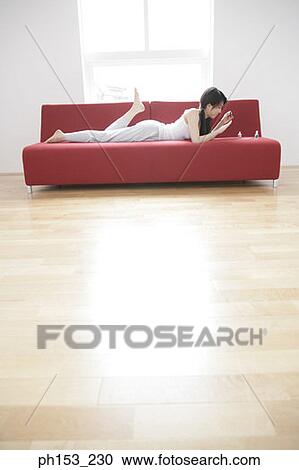 Stock Image - Side profile of a young woman lying on a couch and applying nail polish on her fingernails. Fotosearch