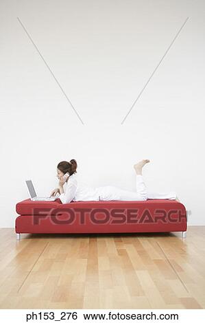 Side profile of a young woman lying on a couch and using a laptop while talking on a mobile phone View Large Photo Image Stock Photograph - Side profile of a young woman lying on a couch and using a laptop while talking on a mobile phone. Fotosearch