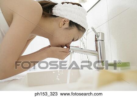 Stock Image - Side profile of a young woman washing her face over a bathroom sink. Fotosearch