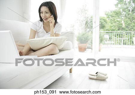 Young woman sitting on a couch and using a laptop while talking on a mobile phone View Large Photo Image Stock Image - Young woman sitting on a couch and using a laptop while talking on a mobile phone. Fotosearch