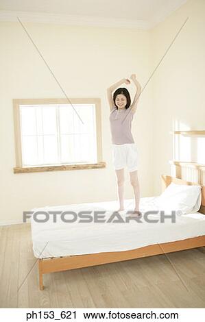 Stock Image - Young woman standing on the bed and stretching her arms. Fotosearch