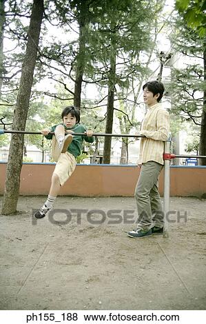 Stock Photo - Boy hanging on monkey bars with his father standing beside him. Fotosearch