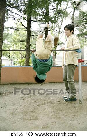 Stock Photo - Boy hanging on monkey bars with his father standing beside him. Fotosearch