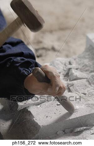 Stock Image - Close-up of a person's hand using a chisel and a hammer on a stone. Fotosearch