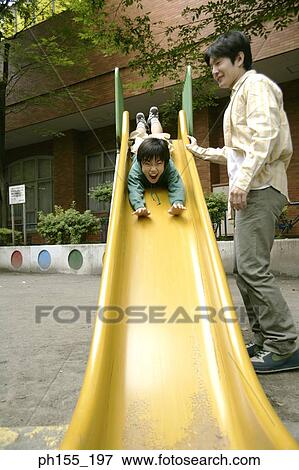Stock Photo - Low angle view of a boy sliding on a slide with his father standing beside him. Fotosearch