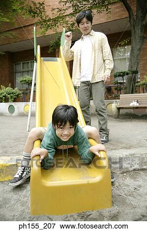 Stock Photo - Low angle view of a boy sliding on a slide with his father standing beside him. Fotosearch