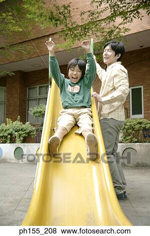 Stock Photo - Low angle view of a boy sliding on a slide with his father standing beside him. Fotosearch