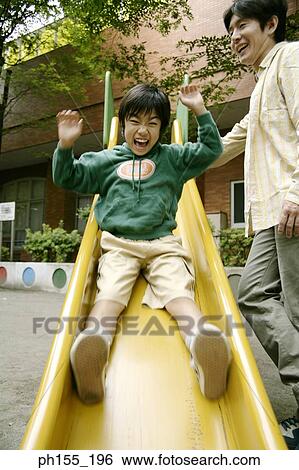 Stock Photograph - Low angle view of a boy sliding on a slide with his father standing beside him. Fotosearch