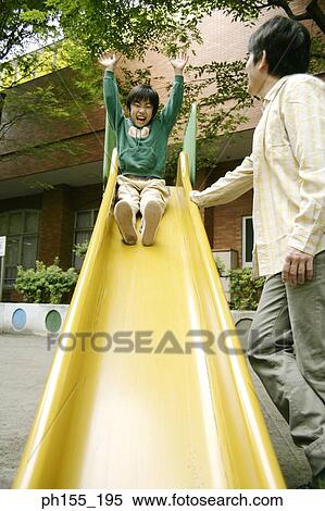 Stock Photography - Low angle view of a boy sliding on a slide with his father standing beside him. Fotosearch