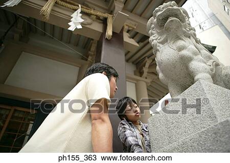 Stock Image - Low angle view of a mid adult man and his son standing near a statue of a lion. Fotosearch