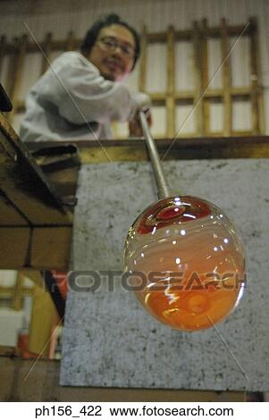 Stock Image - Low angle view of a mid adult man blowing molten glass with a blow pipe. Fotosearch