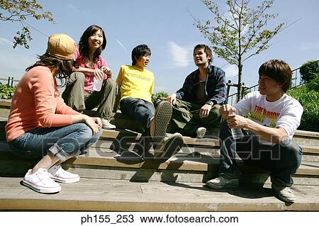 Stock Image - Low angle view of three young men with two young women sitting on steps and smiling. Fotosearch