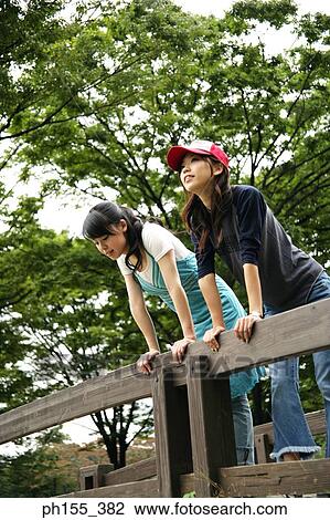 Stock Image - Low angle view of two young women leaning against a wood railing. Fotosearch