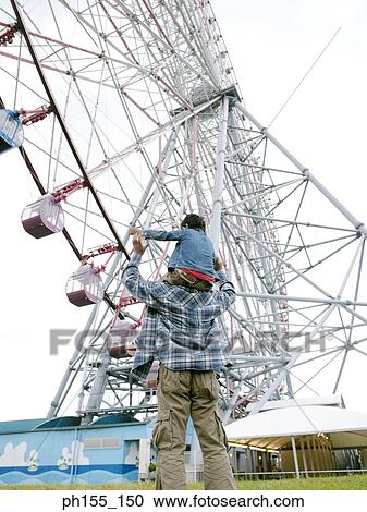 Stock Image - Rear view of a man carrying his child on his shoulders near a ferris wheel. Fotosearch