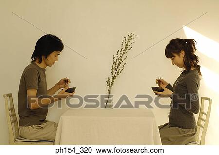 Stock Image - Side profile of a young couple holding bowls and chopsticks. Fotosearch