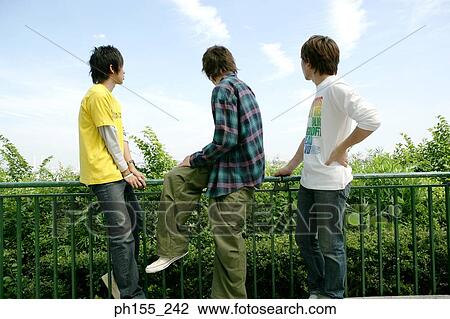 Side profile of two men sitting on a railing with another man standing beside them View Large Photo Image Stock Image - Side profile of two men sitting on a railing with another man standing beside them. Fotosearch