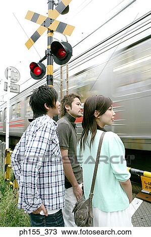 Stock Image - Side profile of two young men and a young woman standing at a railway crossing. Fotosearch