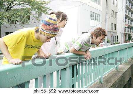 Stock Image - Two young men and a young woman leaning over a railing and looking down. Fotosearch