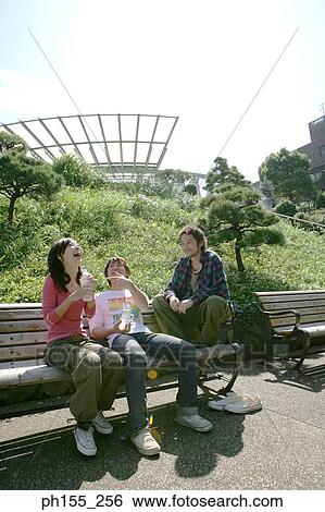 Two young men and a young woman sitting on a park bench and smiling View Large Photo Image Stock Photograph - Two young men and a young woman sitting on a park bench and smiling. Fotosearch