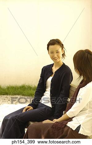 Stock Photo - Two young women sitting on a bench and smiling. Fotosearch