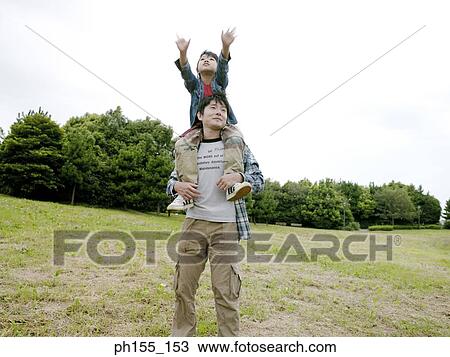 Young man carrying his son on his shoulders and standing in a field View Large Photo Image Stock Image - Young man carrying his son on his shoulders and standing in a field. Fotosearch