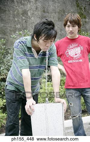 Young man drinking water from a drinking fountain with his friend standing beside him View Large Photo Image Stock Photo - Young man drinking water from a drinking fountain with his friend standing beside him. Fotosearch