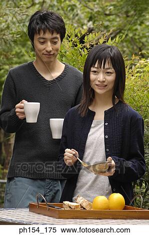 Stock Photography - Young woman holding a bowl and a fork with a young man standing behind her holding coffee cups. Fotosearch