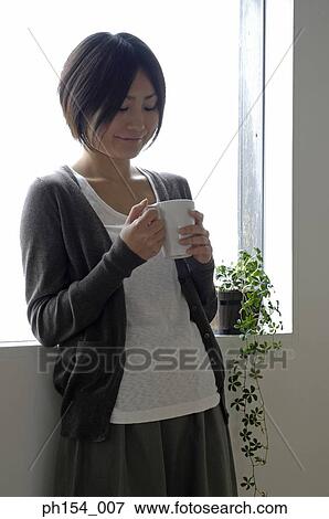 Stock Photo - Young woman holding a coffee cup and leaning against a window. Fotosearch