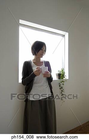 Young woman holding a coffee cup and standing by a window View Large Photo Image Stock Photo - Young woman holding a coffee cup and standing by a window. Fotosearch