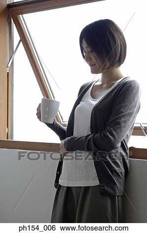 Young woman holding a coffee cup and leaning against a window View Large Photo Image Stock Photograph - Young woman holding a coffee cup and leaning against a window. Fotosearch