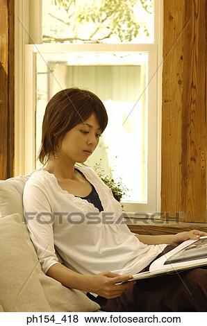 Young woman sitting on a couch and looking at a magazine View Large Photo Image Stock Photo - Young woman sitting on a couch and looking at a magazine. Fotosearch