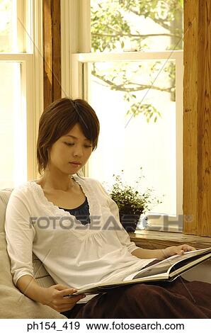 Young woman sitting on a couch and looking at a magazine View Large Photo Image Stock Photo - Young woman sitting on a couch and looking at a magazine. Fotosearch