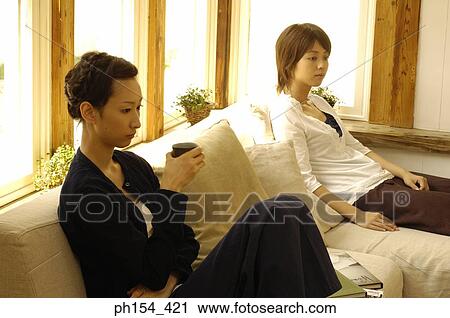 Young woman sitting on a couch holding a tea cup with another young woman sitting beside her View Large Photo Image Stock Image - Young woman sitting on a couch holding a tea cup with another young woman sitting beside her. Fotosearch