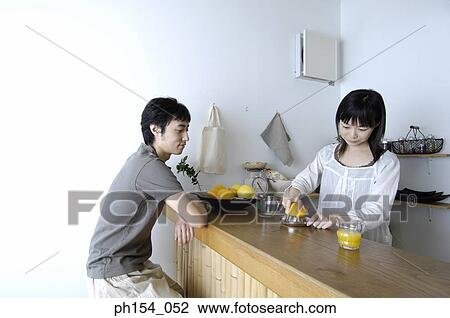Stock Image - Young woman squeezing an orange in a juicer with a young man sitting in front of her. Fotosearch