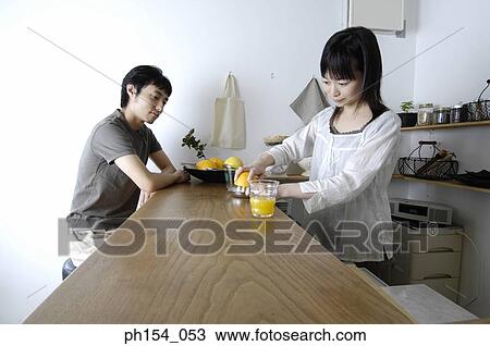 Stock Image - Young woman squeezing an orange in a juicer with a young man sitting in front of her. Fotosearch