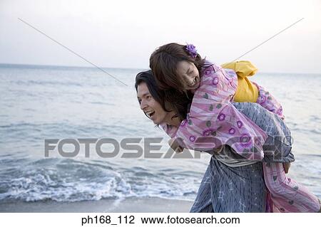 Stock Image - Man walking along the beach with woman on his back. Fotosearch