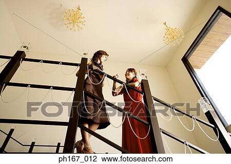 Stock Image - Two women in evening gowns, standing on stairs, low angle. Fotosearch