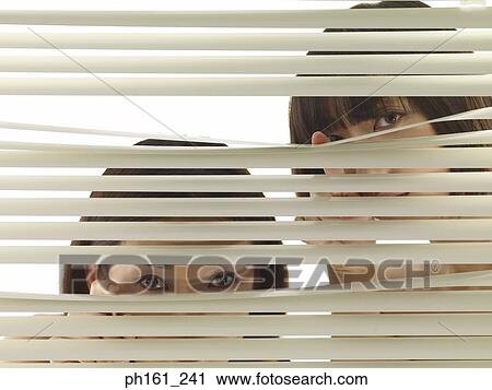 Stock Image - Two women peering through blinds, close-up. Fotosearch