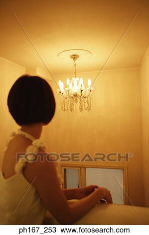 Stock Image - Woman leaning against wall, looking at chandelier, rear view. Fotosearch