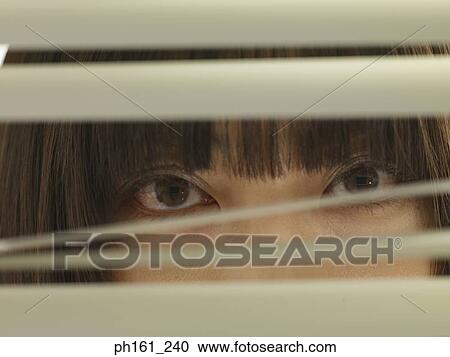 Stock Image - Woman peering through blinds, close-up. Fotosearch