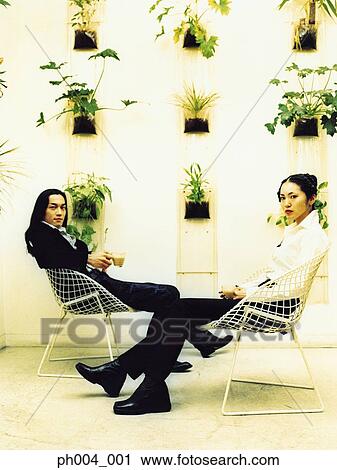 Stock Image - Young couple sitting on wire-framed chairs, potted plants decorated wall background. Fotosearch