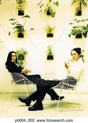 Stock Image - Young couple sitting on wire-framed chairs, potted plants decorated wall background. Fotosearch