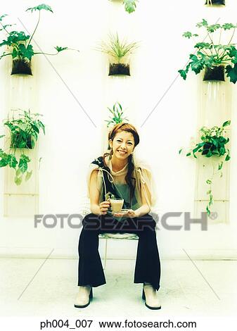 Stock Photo - Young woman sitting holding a cup of beverage, potted plants decorated wall background. Fotosearch