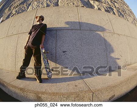 Picture - Young male standing against wall holding skateboard . Fotosearch