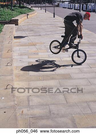 Young man performing bike stunt on pavement View Large Photo Image Stock Photograph - Young man performing bike stunt on pavement. Fotosearch